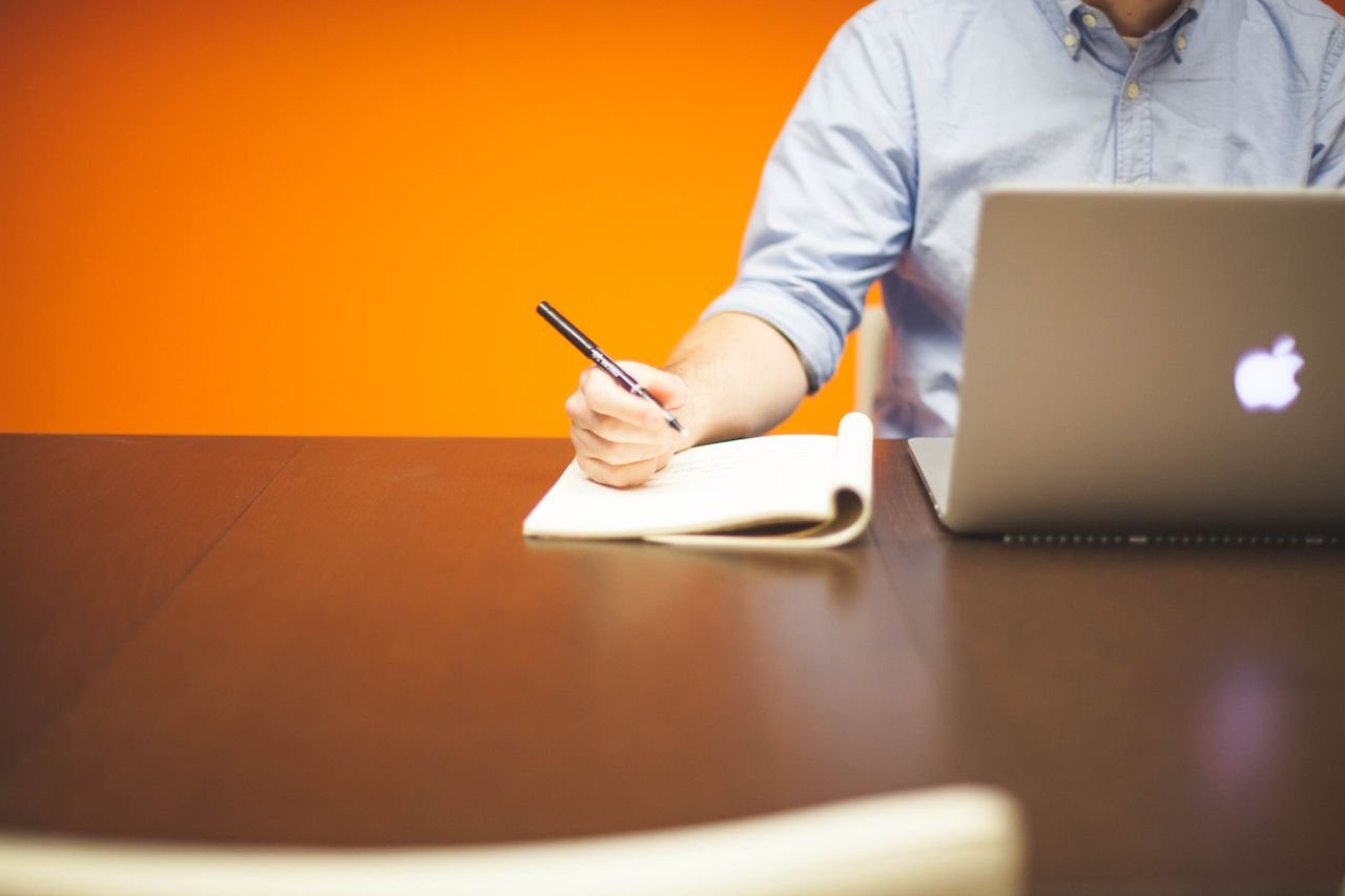 Perfil para teletrabajo técnico, soporte remoto y sistemas Adult writing notes in a notebook beside a laptop on a wooden desk with orange background.